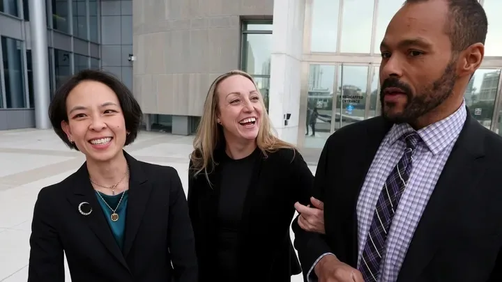 Three people stand outside a modern building; the woman on the left smiles, the woman in the middle laughs, and the man on…