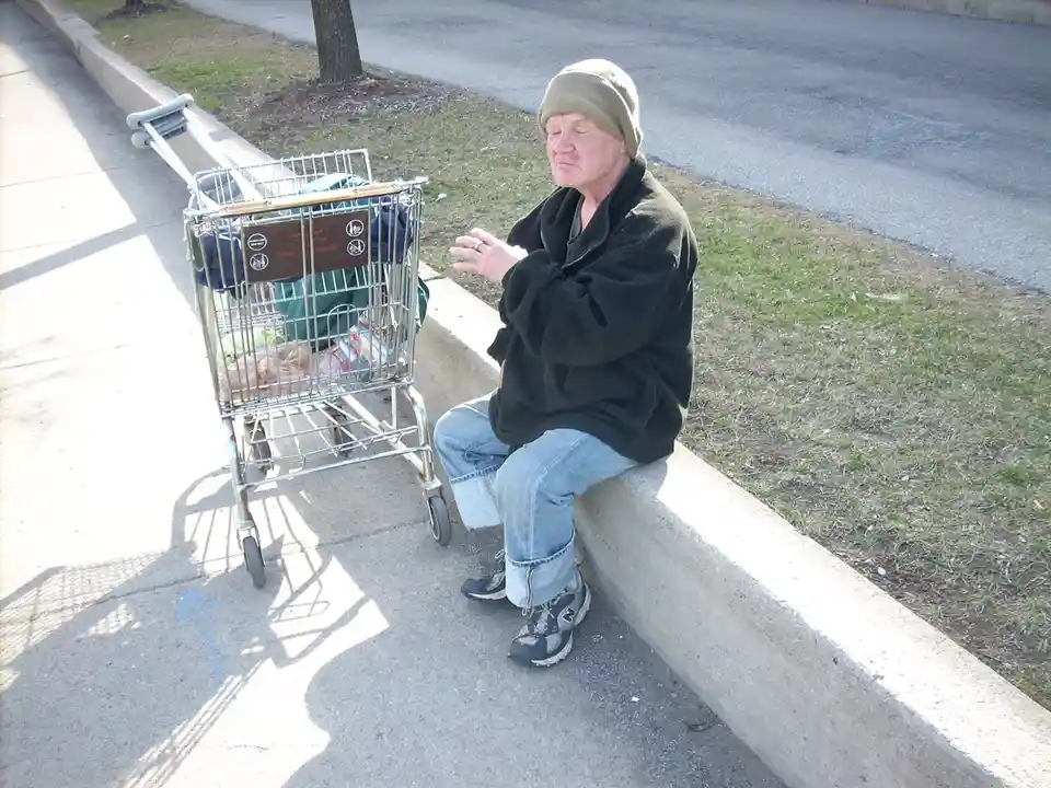 An older man wearing a beanie, black jacket, and jeans sits on a concrete curb next to a shopping cart filled with…