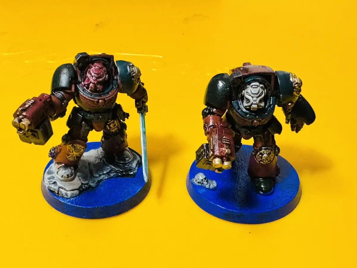 Two painted Warhammer 40k Space Marine miniatures stand side-by-side on blue circular bases against a bright yellow…