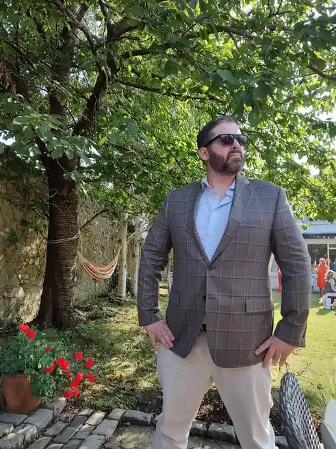 A man with a beard and sunglasses stands outdoors in a garden, wearing a plaid blazer and light pants, hands on hips…