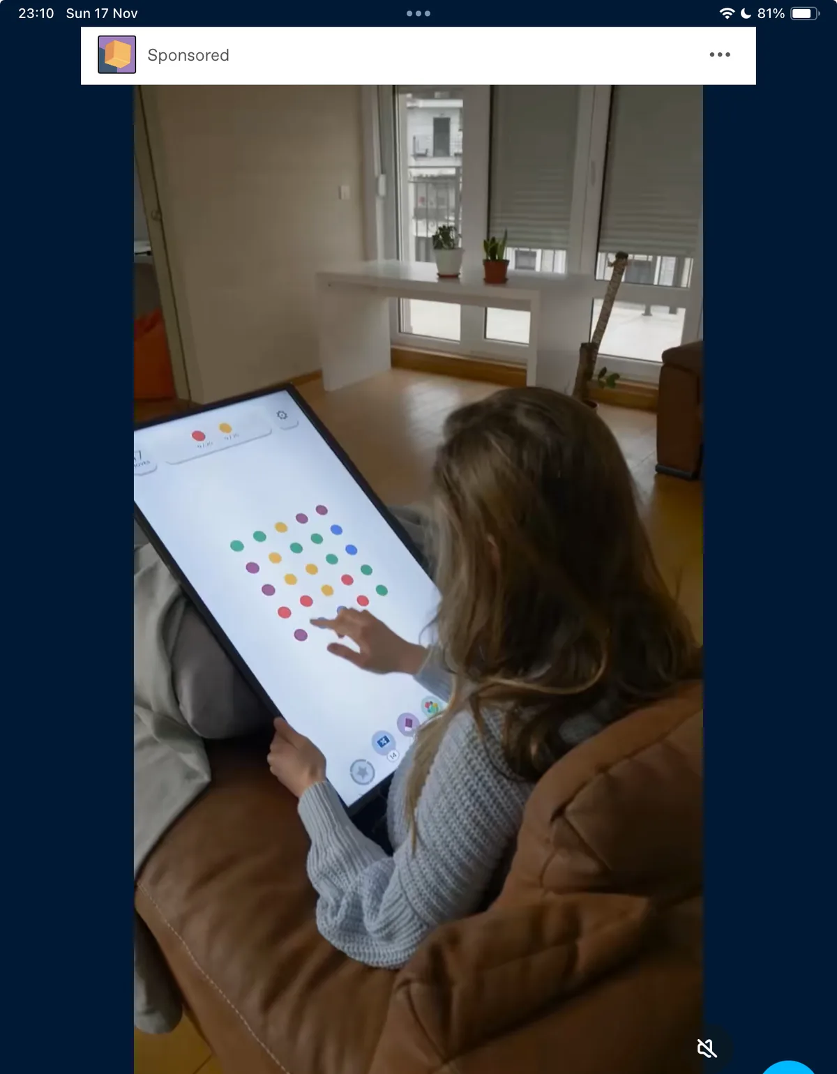 A person is sitting on a couch, holding a large tablet displaying a colorful dot-matching game. The screen shows a grid of…