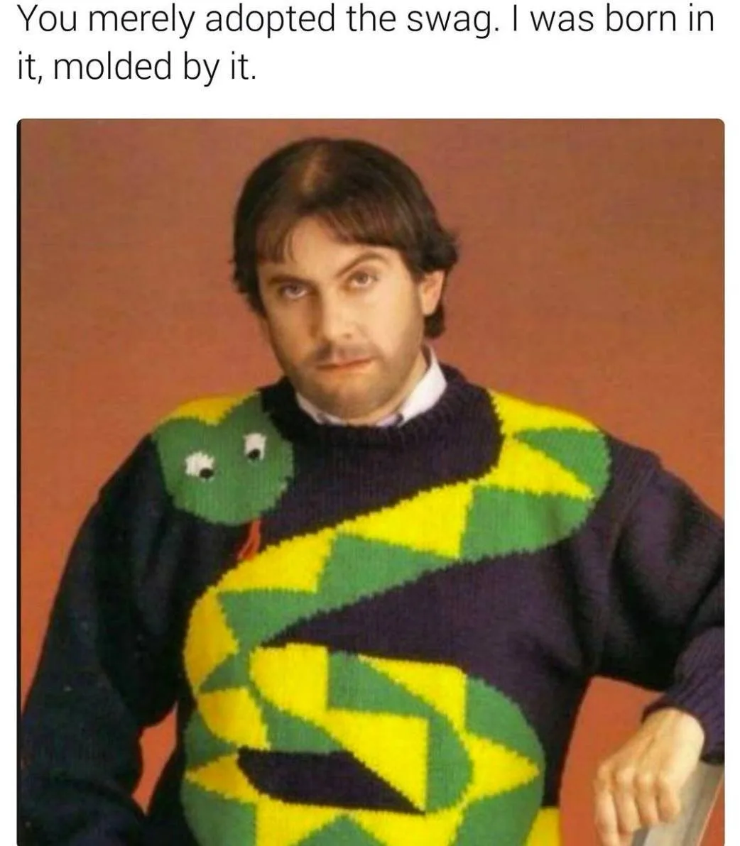 A man with dark hair and a mustache stares directly at the camera with a smug expression, wearing a dark purple sweater…