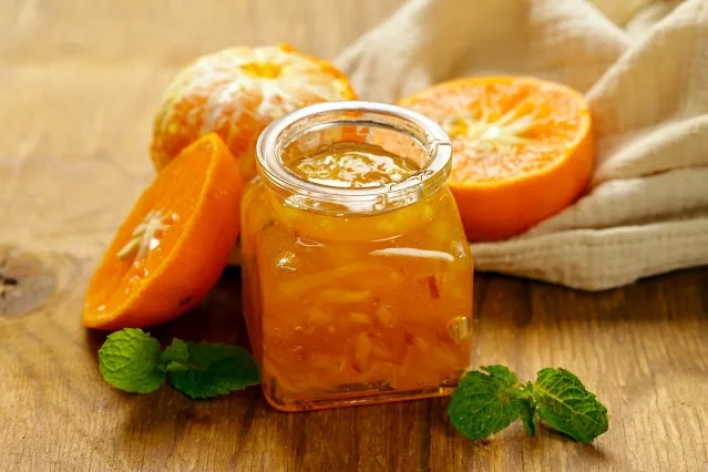 A glass jar of orange marmalade sits on a wooden surface, surrounded by fresh orange halves, a peeled orange, and sprigs of…