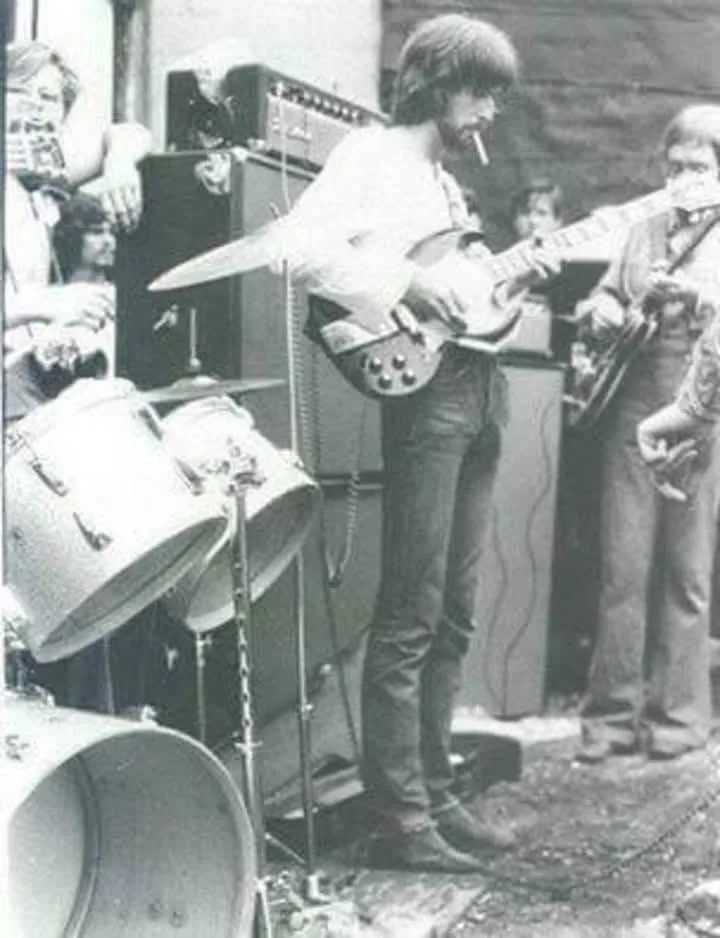 A grainy black-and-white photo of a 1960s rock band performing outdoors, with a guitarist in the center smoking a cigarette…