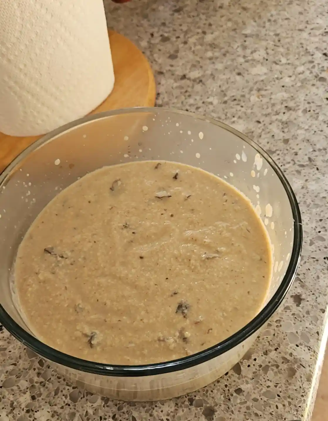 A clear glass bowl sits on a speckled countertop, filled with a thick, beige batter speckled with dark flecks, likely…