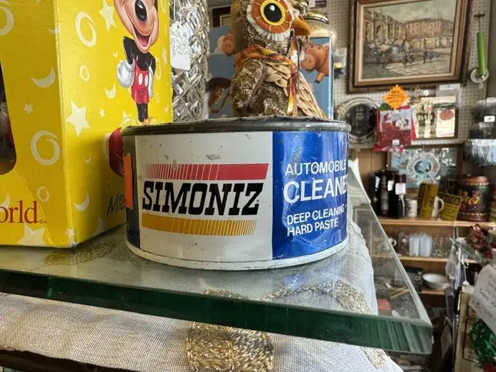 A vintage, weathered metal can of Simoniz Automobile Cleaner, labeled 'Deep Cleaning Hard Paste,' sits on a glass shelf in a…
