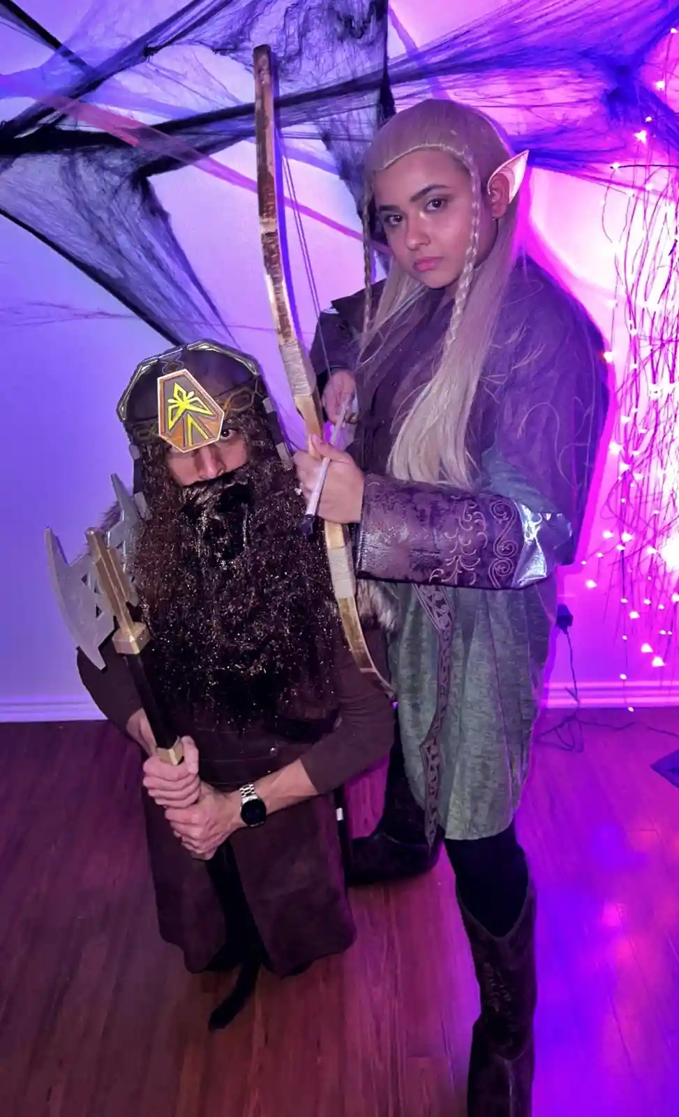 Two people in fantasy costumes pose indoors under purple lighting; one wears a dwarf helmet and holds an axe, while the…