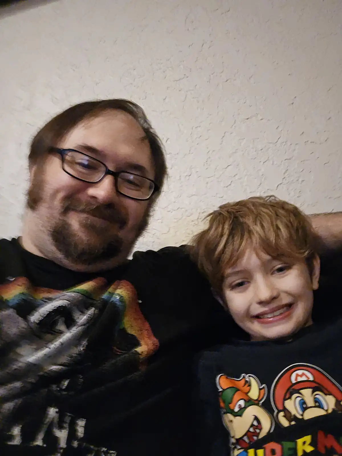 A man with glasses and a beard smiles while embracing a young boy with reddish hair who is wearing a Mario-themed t-shirt…