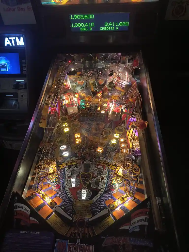 A brightly lit pinball machine with a medieval theme, featuring a large central crest and glowing targets, sits next to an…