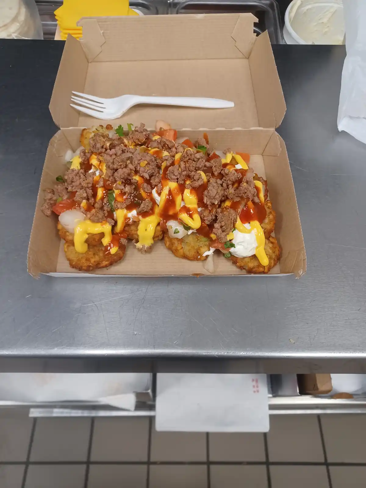A cardboard food box sits open on a stainless steel kitchen counter, revealing a serving of loaded tater tots topped with…