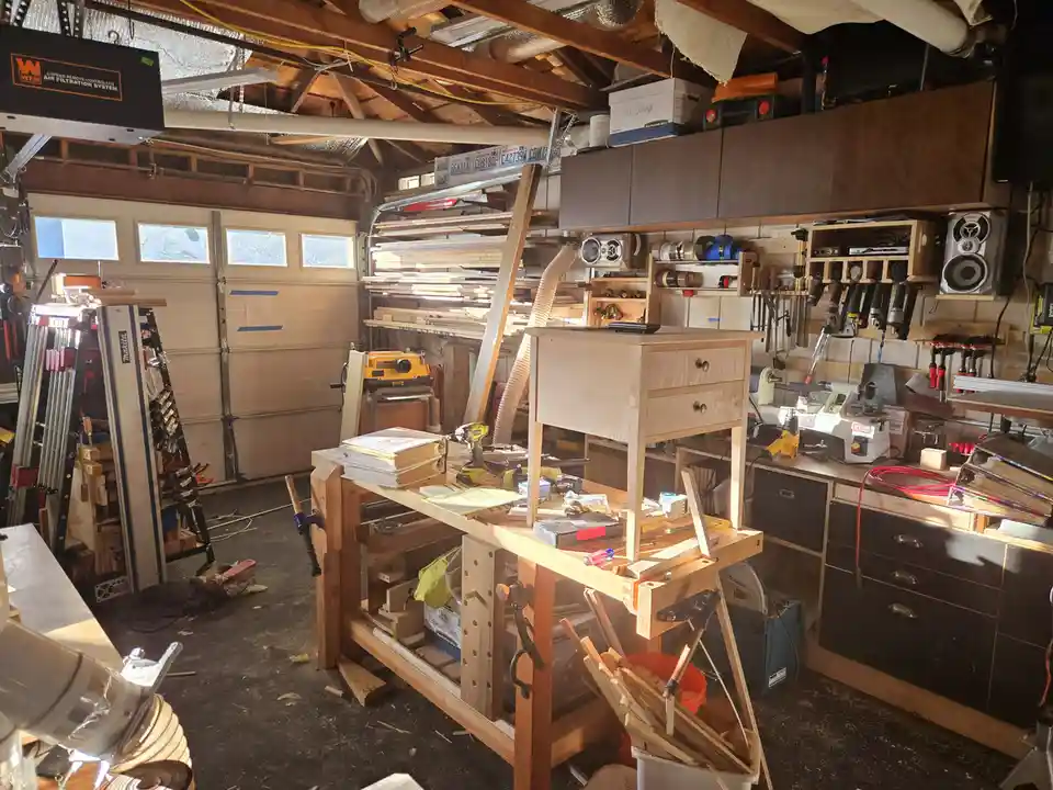 A cluttered woodworking workshop filled with tools, wood, and equipment, featuring a small unfinished wooden dresser sitting…