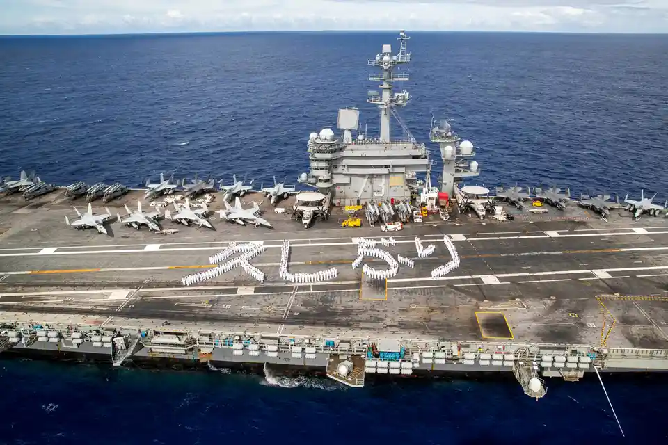An aerial view of an aircraft carrier at sea, with numerous fighter jets parked on its deck. A group of sailors in white…