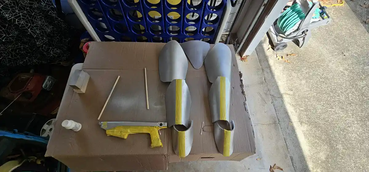 A cardboard box outdoors holds several silver-painted shovel blade pieces taped with yellow tape, alongside a small spray…