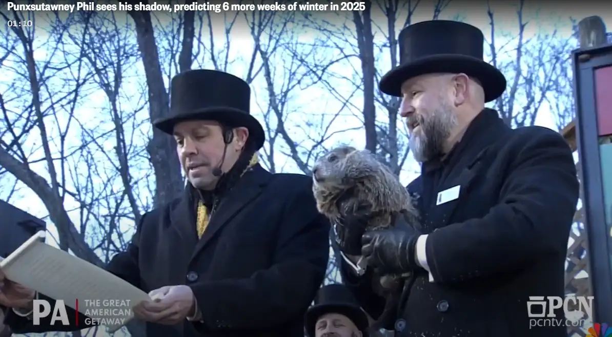 Two men in black top hats and coats stand outdoors in winter, one holding a live groundhog while the other reads from a…