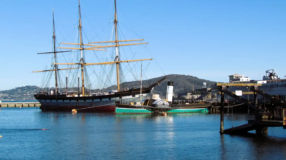 A large, historic three-masted sailing ship is docked in a harbor next to a smaller steamboat, with hills and a pier visible…