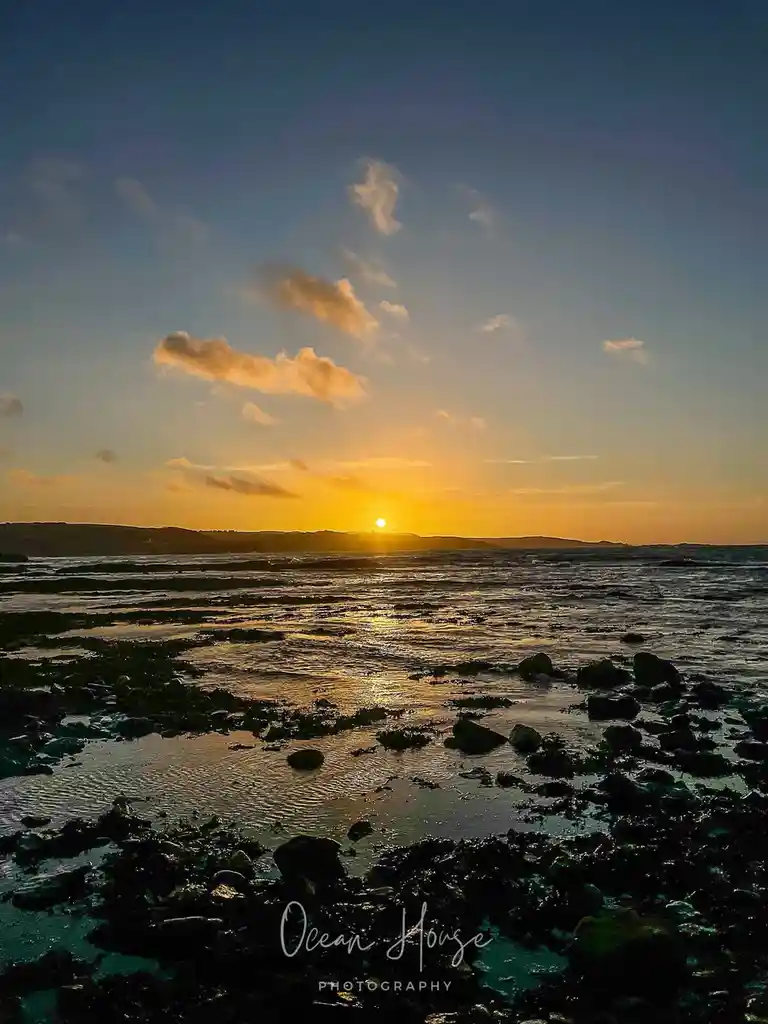 A serene sunset photograph over a rocky shoreline with the sun low on the horizon, casting golden light across the water and…