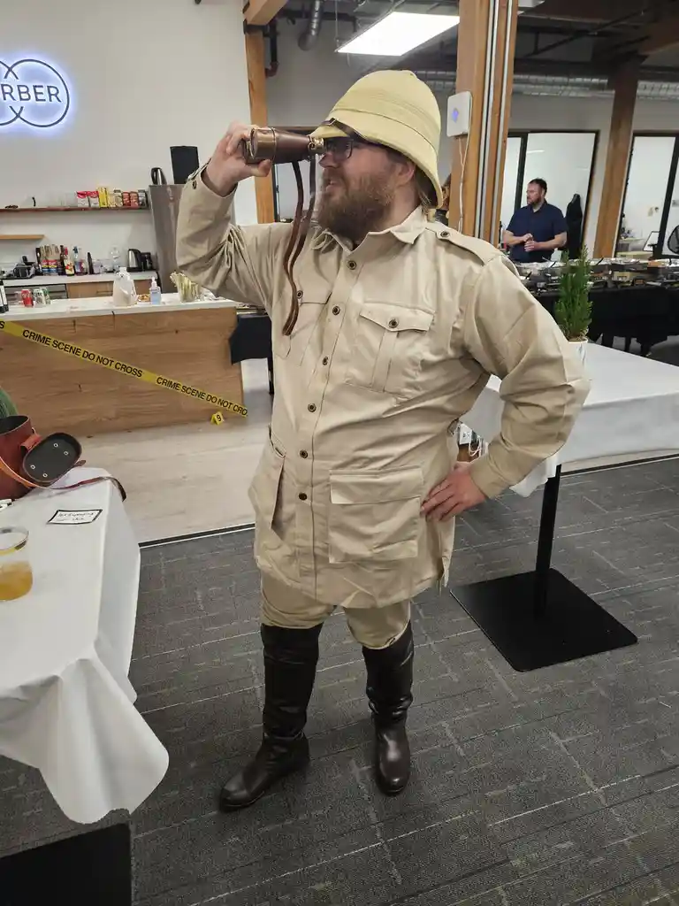 A man in a beige safari outfit with a pith helmet and binoculars stands in an office, looking through the binoculars with…