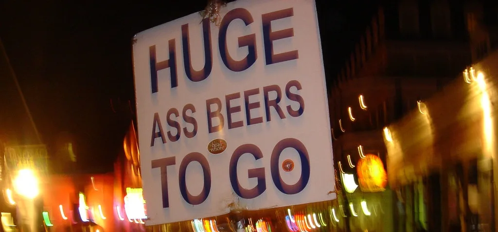 A blurry nighttime photo shows a white sign with large, bold, dark brown text reading 'HUGE ASS BEERS TO GO'. The sign is…