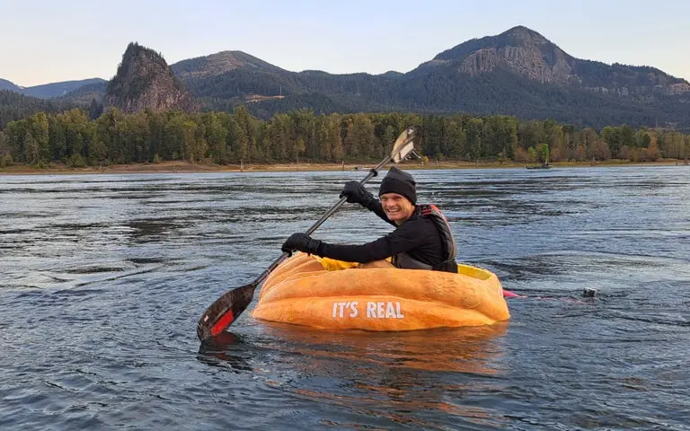 A person wearing a black beanie and jacket paddles a large, orange, pumpkin-shaped kayak on a river with mountains in the…