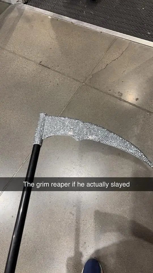A close-up photo shows a scythe with a black handle and a blade entirely covered in sparkling rhinestones, lying on a…