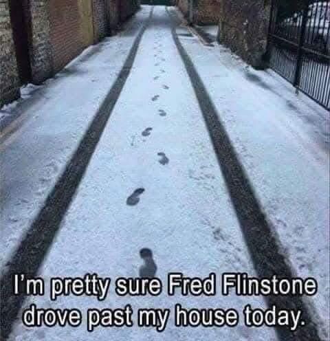 A snowy alleyway with two parallel tire tracks and a single trail of human footprints leading away from the viewer. The…