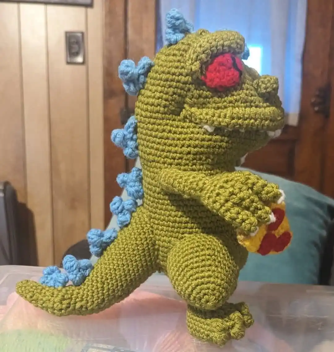 A handmade, crocheted green Godzilla toy with blue dorsal fins and red eyes stands on a clear plastic surface. It holds a…