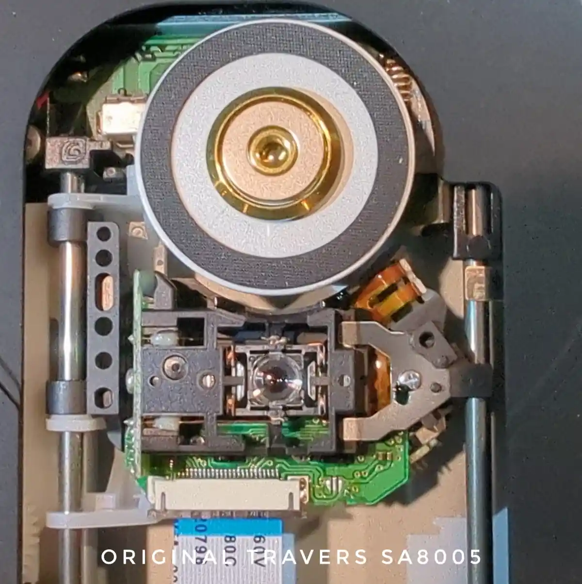 A close-up photo of the internal mechanism of a CD or DVD drive, showing its laser lens assembly, spinning disc hub, and…
