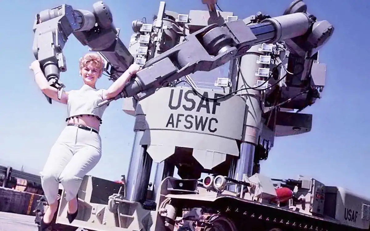 A woman in a light-colored uniform smiles while posing with her hands on the arms of a large, complex military robot labeled…