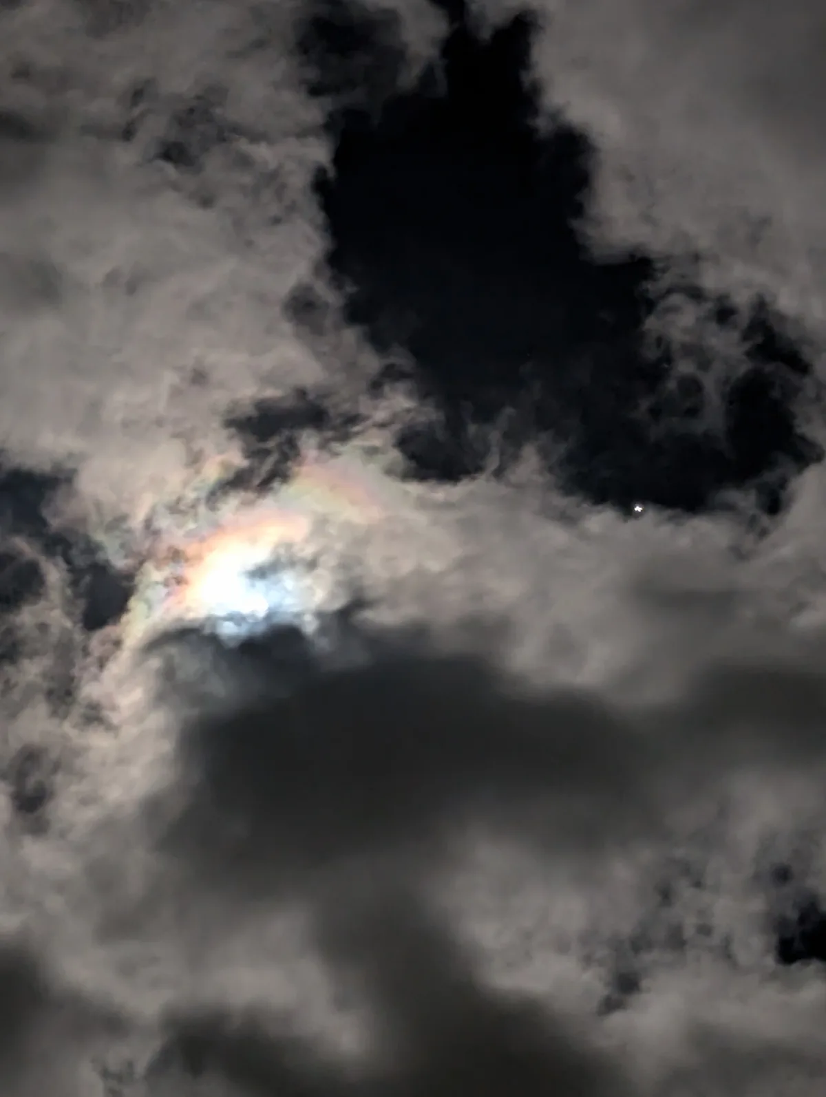 A blurry nighttime photo shows a bright moon partially obscured by dark, textured clouds, with a faint rainbow-like halo…