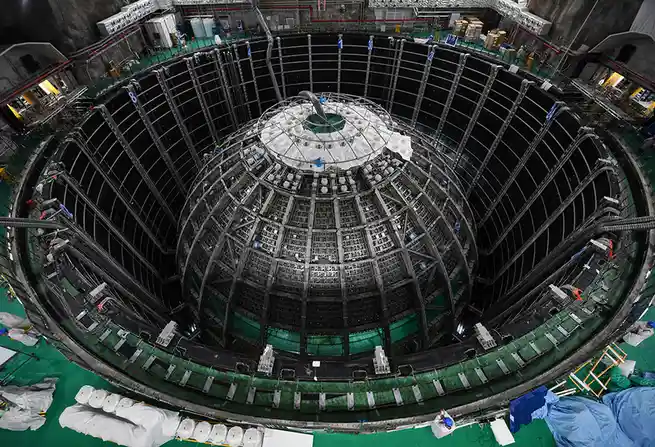 An overhead view of a massive, circular, industrial structure with a complex lattice of metal beams and machinery…
