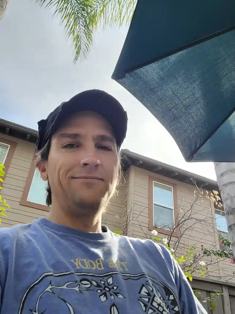 A man wearing a backward black cap and a blue t-shirt with 'THE BODY' printed on it takes a selfie outdoors under a large…