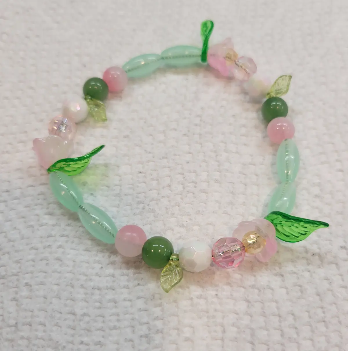 A handmade beaded bracelet with pastel pink, mint green, and white beads, interspersed with small glass flower and leaf…