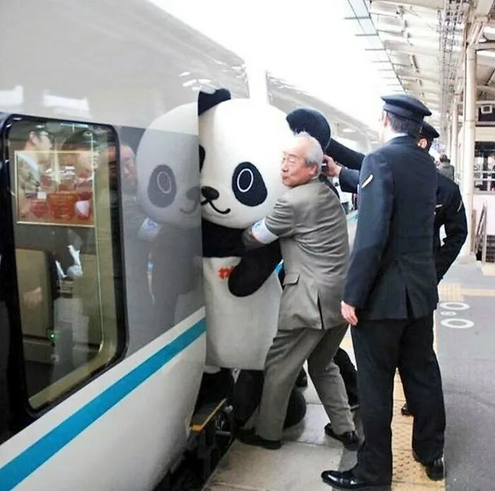An elderly man in a grey suit is being physically assisted by uniformed staff to exit a train, while a large panda mascot…