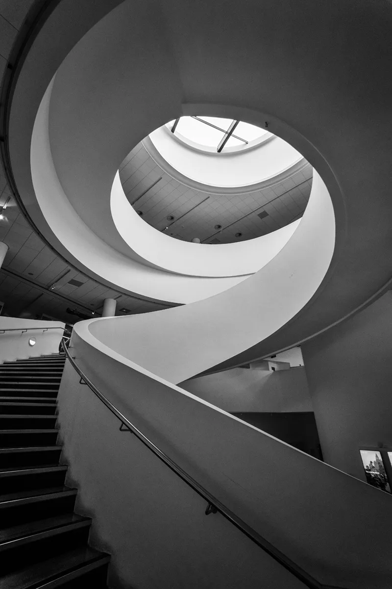 A black-and-white photo shows a sweeping, modern spiral staircase viewed from below, curving upward toward a circular…