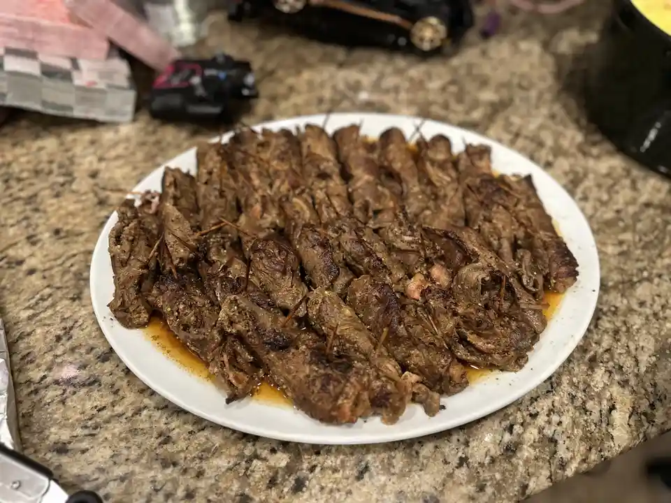 A white plate piled high with numerous grilled meat rolls, each secured with a toothpick, sits on a speckled granite…