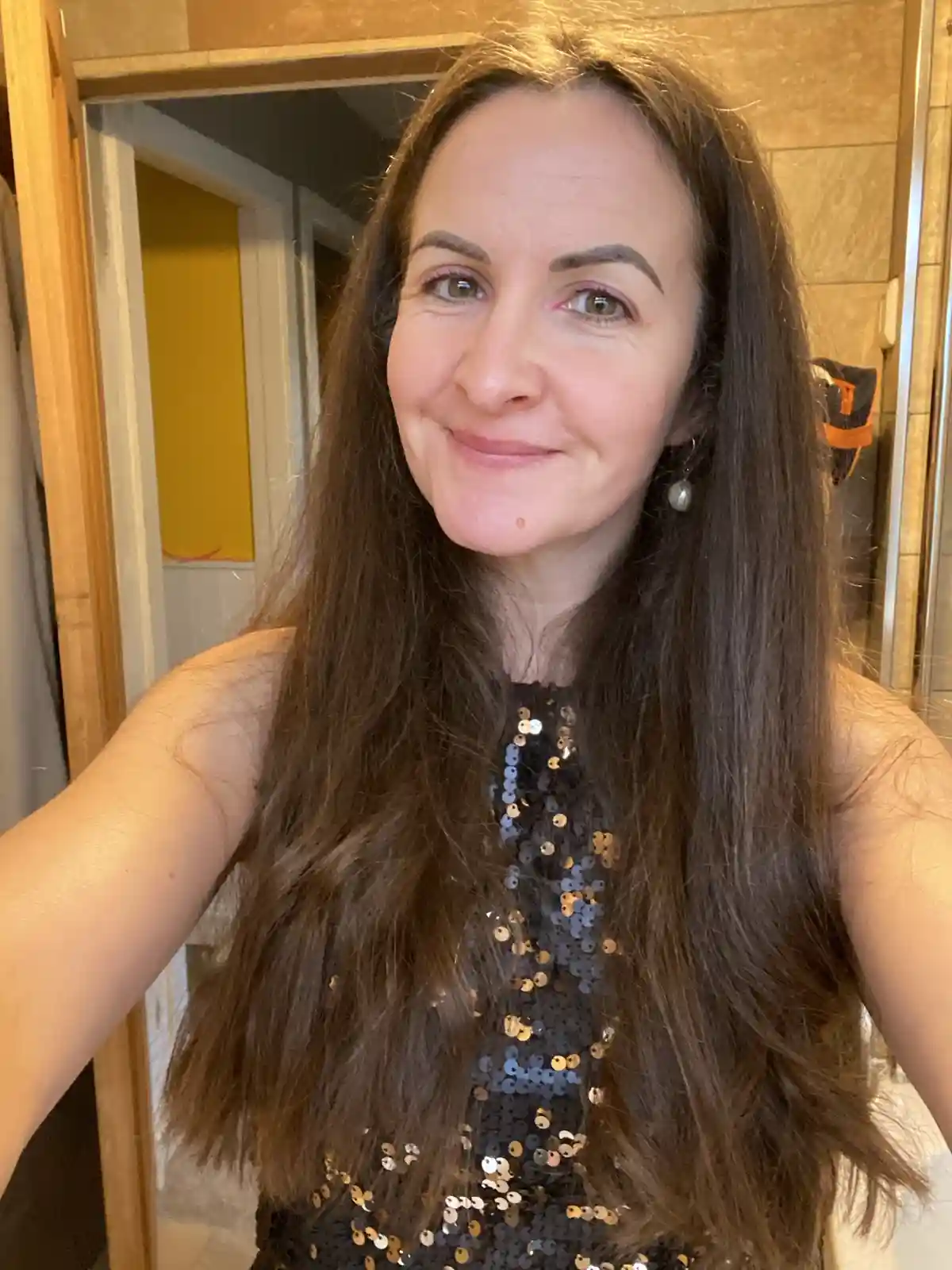 A woman with long brown hair and a sequined top smiles at the camera while taking a selfie in a bathroom, with text overlaid…
