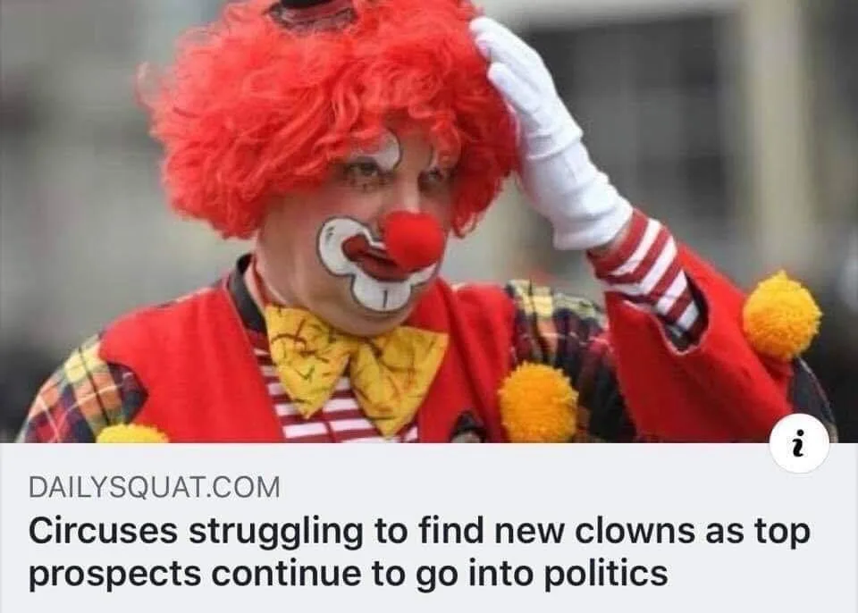 A photo of a male clown with a red wig, red nose, white gloves, and a red vest over a plaid shirt, scratching his head in…