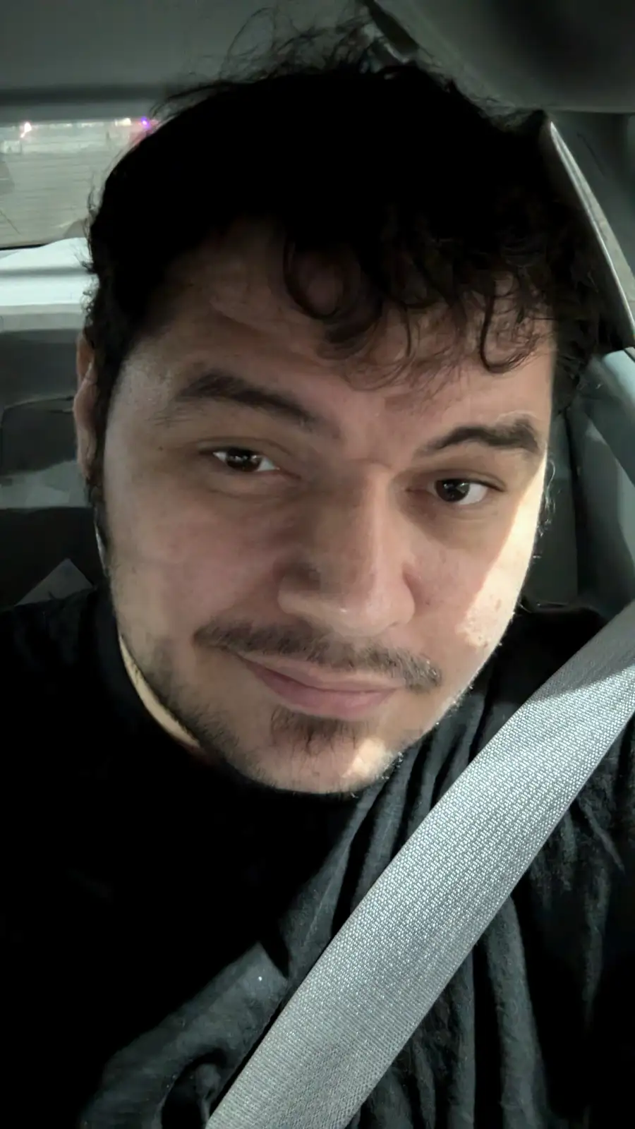 A close-up selfie of a man with dark curly hair and a goatee, wearing a black shirt and a seatbelt, sitting inside a car at…