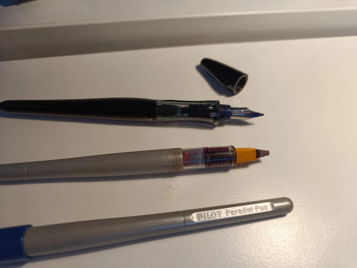Three Pilot pens lie on a white surface: a black fountain pen with its cap off, a grey rollerball pen with a yellow tip and…