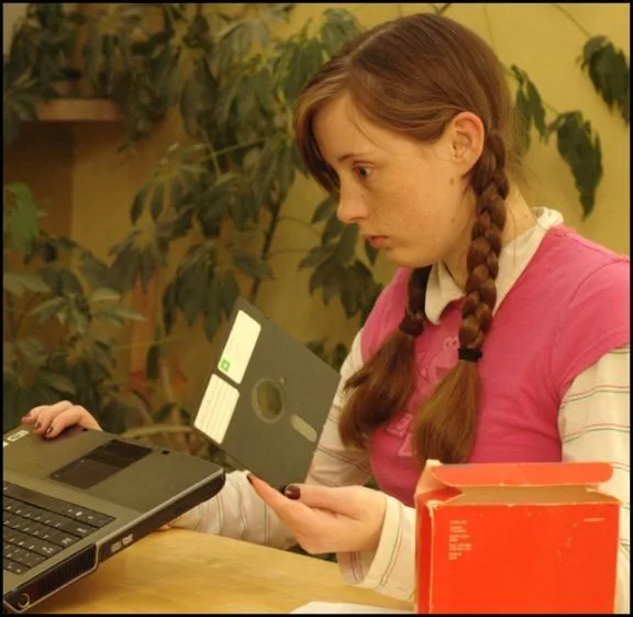A young woman with braided hair sits at a desk holding a floppy disk while looking at a laptop. She has a confused or…