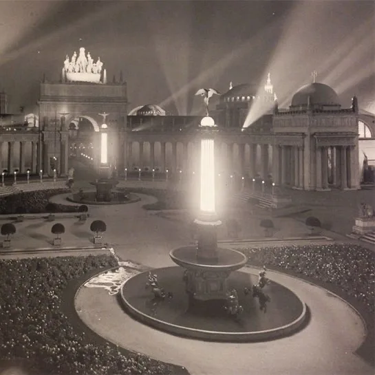 A black-and-white photograph of the illuminated Deutsches Museum in Munich at night, featuring a central fountain with a…