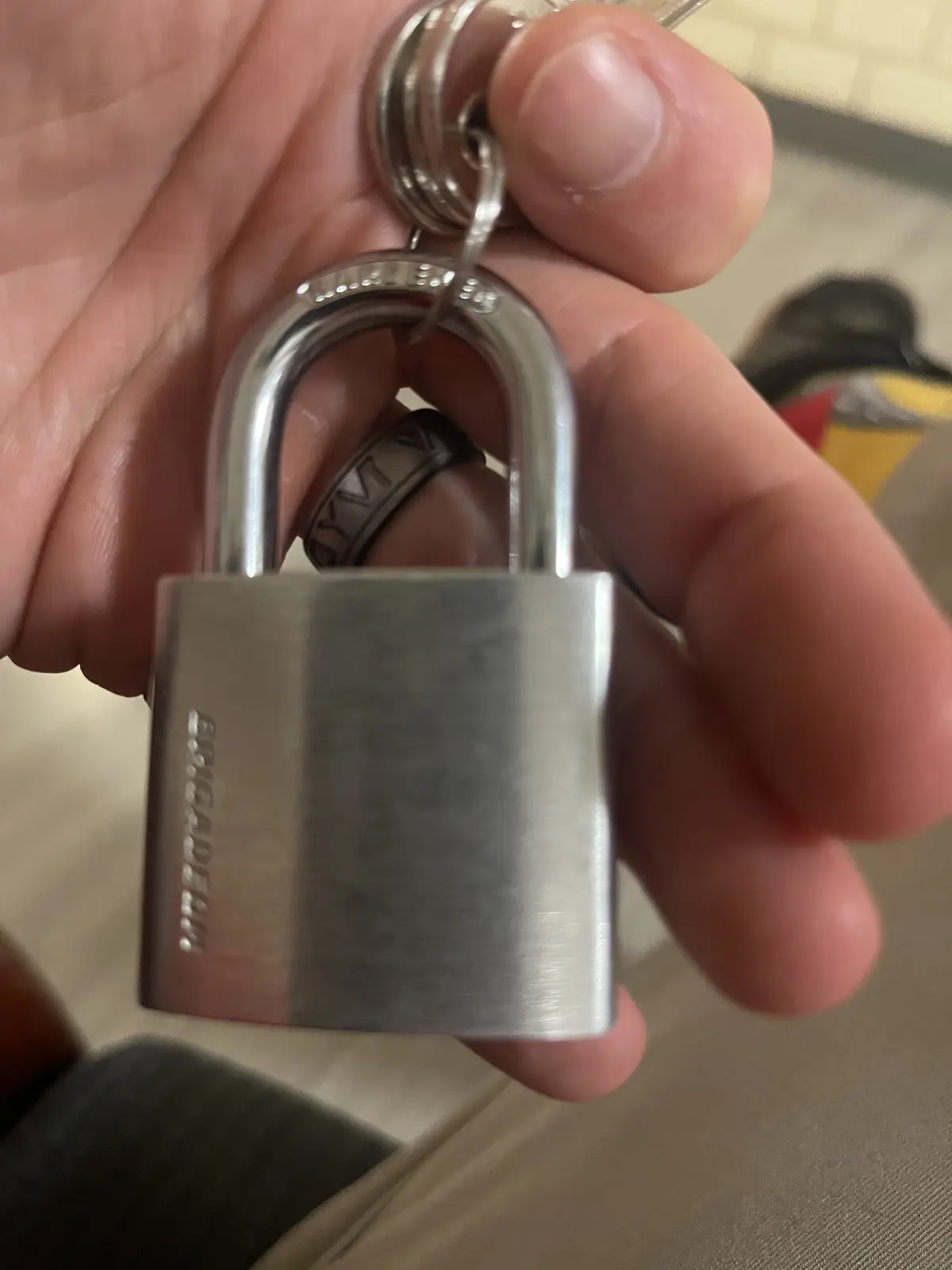 A close-up photo shows a person's hand holding a silver-colored padlock attached to a keychain. The padlock is branded…