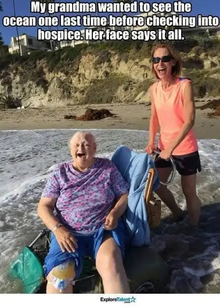 An elderly woman with a joyful, open-mouthed expression sits in a beach wheelchair at the water's edge, waves crashing…