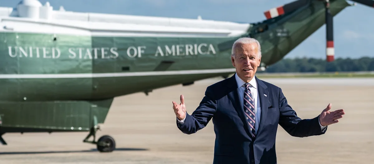 a man in a suit and tie stands with arms outstretched in front of a large green airplane labeled 'UNITED STATES OF AMERICA'…