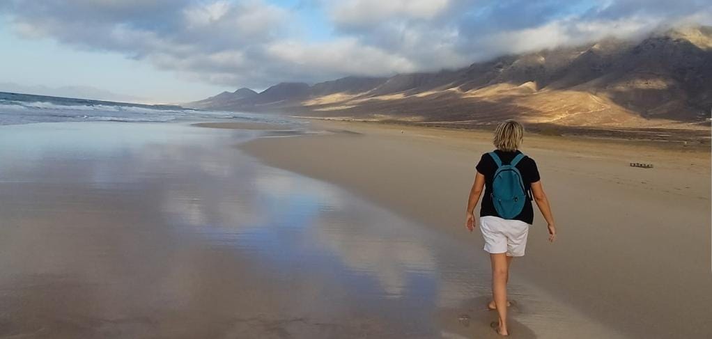 A person with short blonde hair, wearing a black t-shirt, white shorts, and a teal backpack, walks barefoot along a wide…