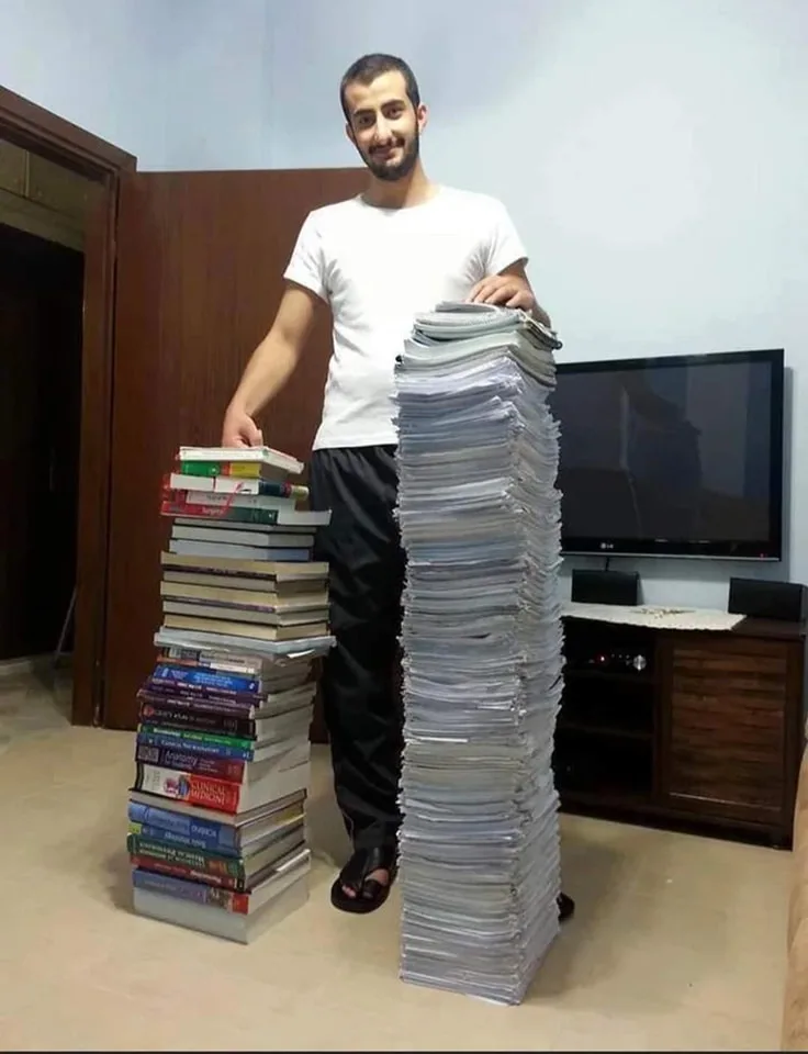 A man in a white t-shirt and black pants stands indoors beside two towering stacks of books and papers, smiling at the…