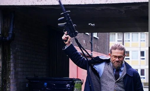 A man with glasses and a beard, wearing a suit jacket and vest, holds a submachine gun raised in the air while standing…