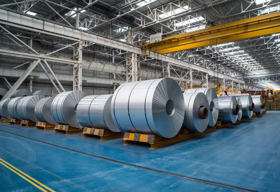 A row of large, shiny metal coils sits on yellow stands inside a vast industrial warehouse with high steel trusses and a…