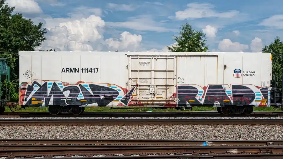 A white Union Pacific boxcar with the ID 'ARMN 111417' and 'BUILDING AMERICA' logo is covered in large, colorful graffiti…