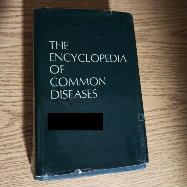 A dark green book titled 'THE ENCYCLOPEDIA OF COMMON DISEASES' sits on a wooden surface, with a black bar obscuring part of…